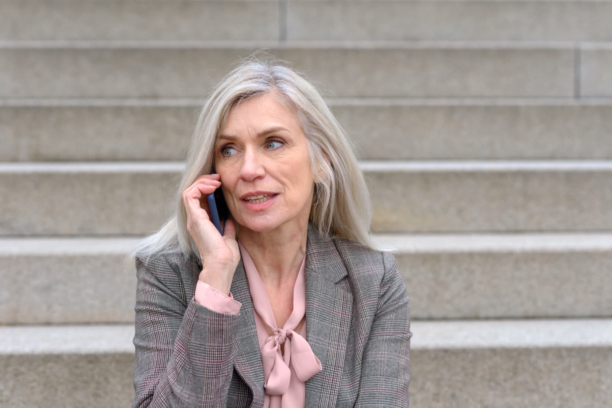 A professional woman sits alone on office steps outside, phone to her ear, her expression tense with concern during a personal call.